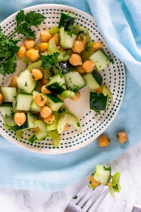 A small white plate with teal dots topped with cucumber chickpea salad with a fork with a bite of salad on it to the side.