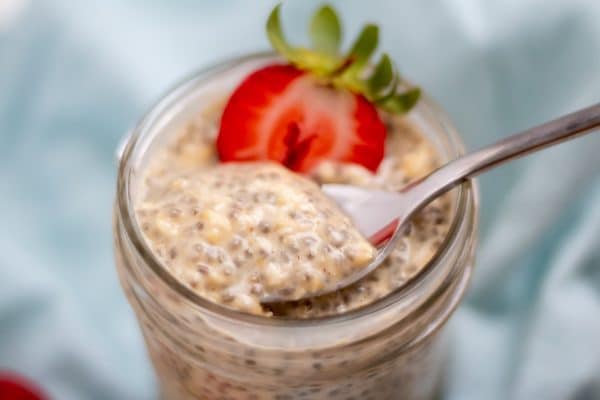 side shot of pale, creamy tan overnight oats with chia seeds with a spoon stirring it and half a strawberry on top