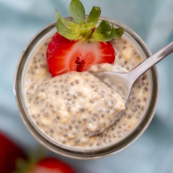 overhead shot of pale, creamy tan overnight oats with chia seeds with a spoon stirring it and half a strawberry on top