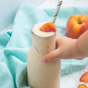 A small child's hand reaching for a glass of creamy banana peach smoothie.