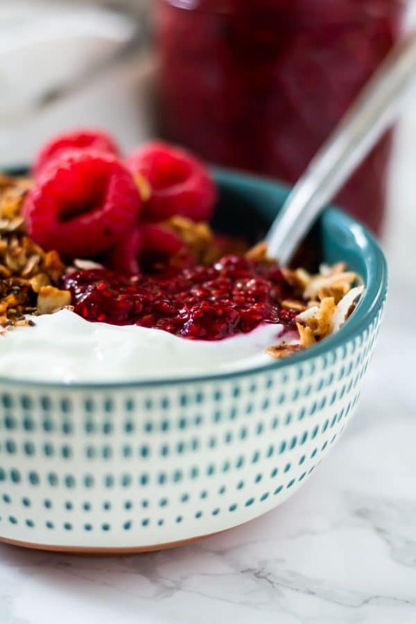 yogurt parfait with raspberries, granola, and raspberry chia jam in a white and teal spotted rustic bowl.