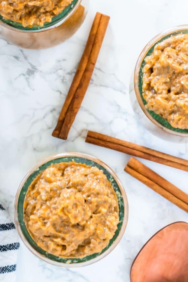 looking down on a marble surface topped with 3 glass jars of pumpkin chia pudding, a copper spoon, and cinnamon sticks