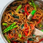 Steel pan of lo mein with bright vegetables and a wooden spoon.