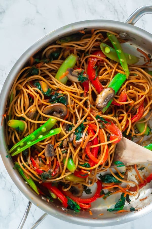 Steel pan of lo mein with bright vegetables and a wooden spoon.