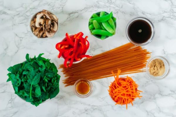 assembled ingredients (spinach, mushrooms, bell pepper, lentil pasta, carrots, sugar snap peas, coconut aminos, brown sugar, and spices) required to make easy, gluten-free lo mein