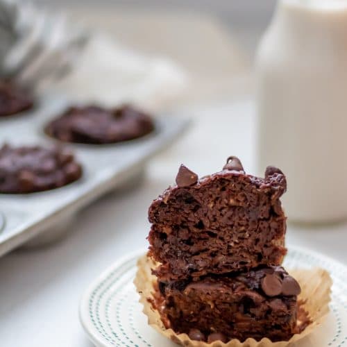 a double chocolate banana muffin cut in half, with the halves stacked on each other and the pan of muffins and bottle of milk in the background.