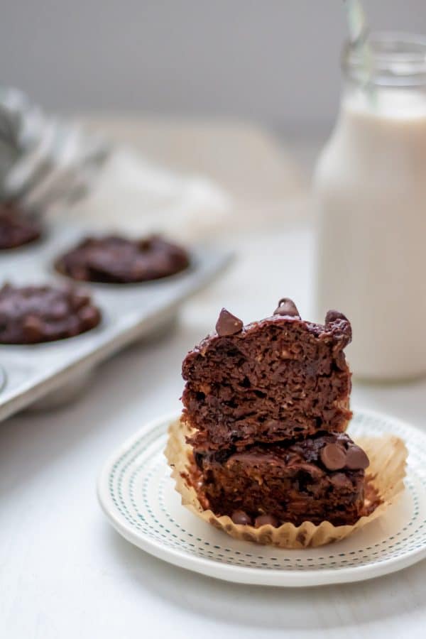 a double chocolate banana muffin cut in half, with the halves stacked on each other and the pan of muffins and bottle of milk in the background.