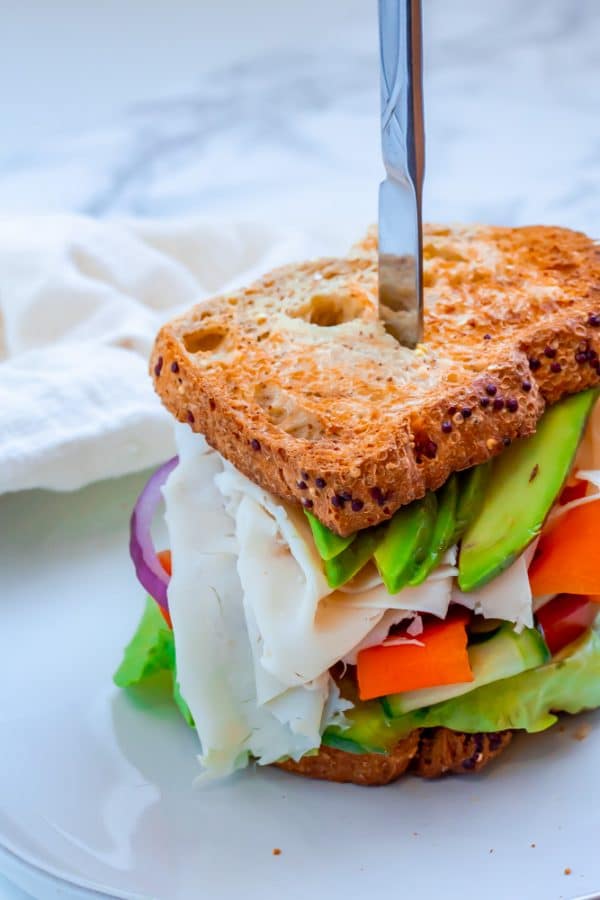 sandwich with deli turkey, avocado, lots of veggies on whole grain bread on a white plate with a butter knife in it.