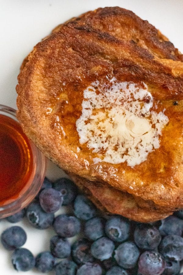 A close up of butter melted on a stack of french toast, next to a cup of maple syrup and fresh blueberries.