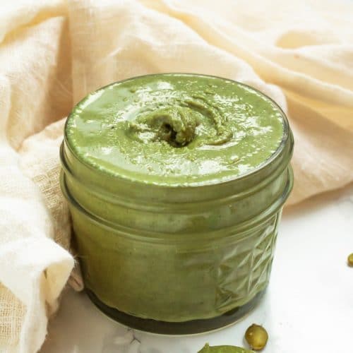 A glass jar of green pumpkin seed butter with pumpkin seeds on a marble surface.