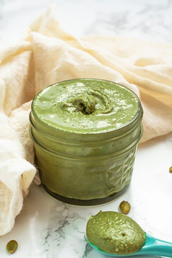 A glass jar of green pumpkin seed butter with pumpkin seeds on a marble surface.