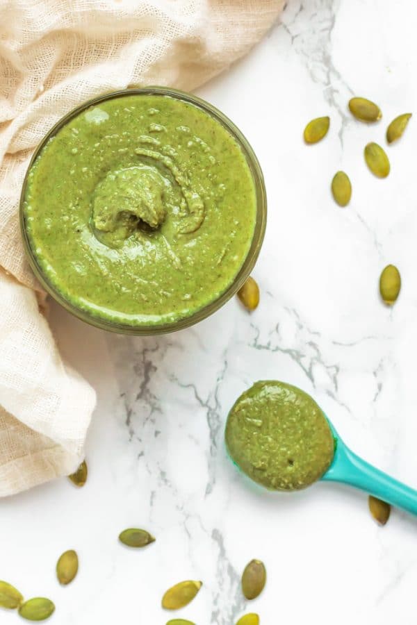 A small glass jar of green pumpkin seed butter with a turquoise spoon on a marble surface.