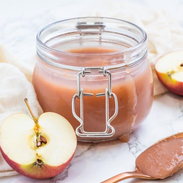 A glass jar of unsweetened applesauce with a spoon of applesauce and apples.