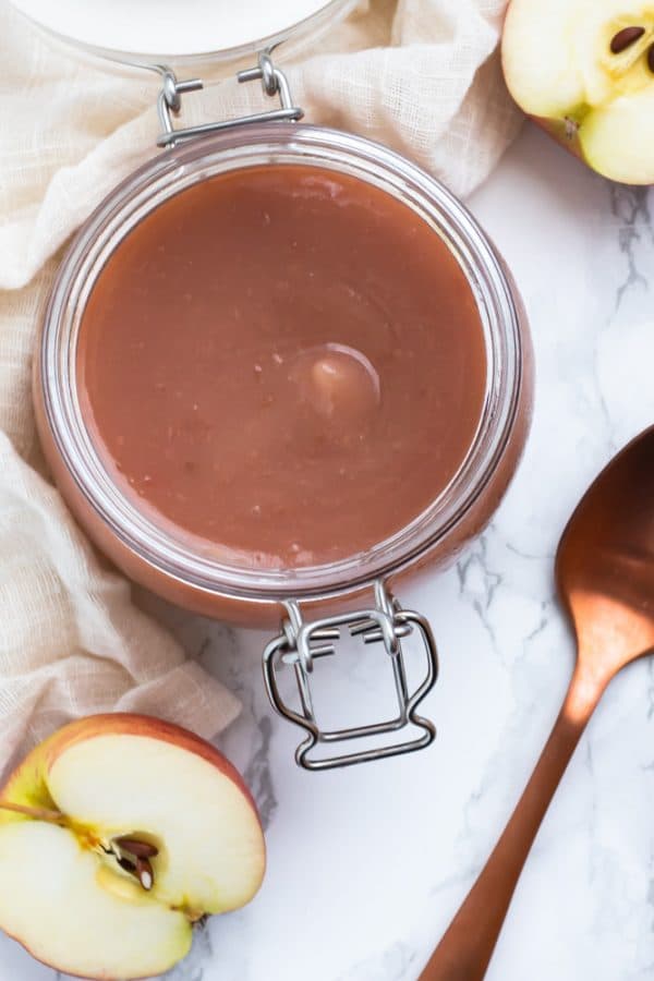 Unsweetened applesauce in a glass jar on a marble surface by a cut apple and copper spoon.
