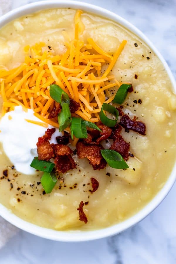 A close up of a white bowl of potato soup topped with bacon, cheese, greek yogurt and green onions.