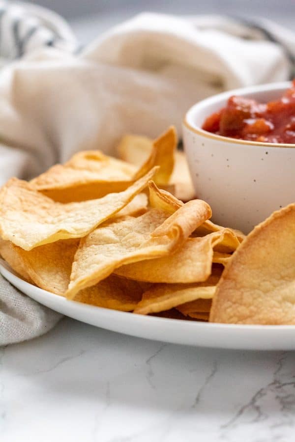 crispy golden brown air fryer tortilla chips on a white plate next to a small white bowl with gold trim full of salsa.