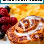 a golden brown cinnamon roll with icing on a plate with fresh berries and scrambled eggs. Text at the top of the image says "air fryer cinnamon rolls"