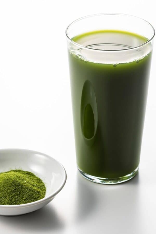 A tall glass of green juice next to a small white bowl of a greens powder supplement.