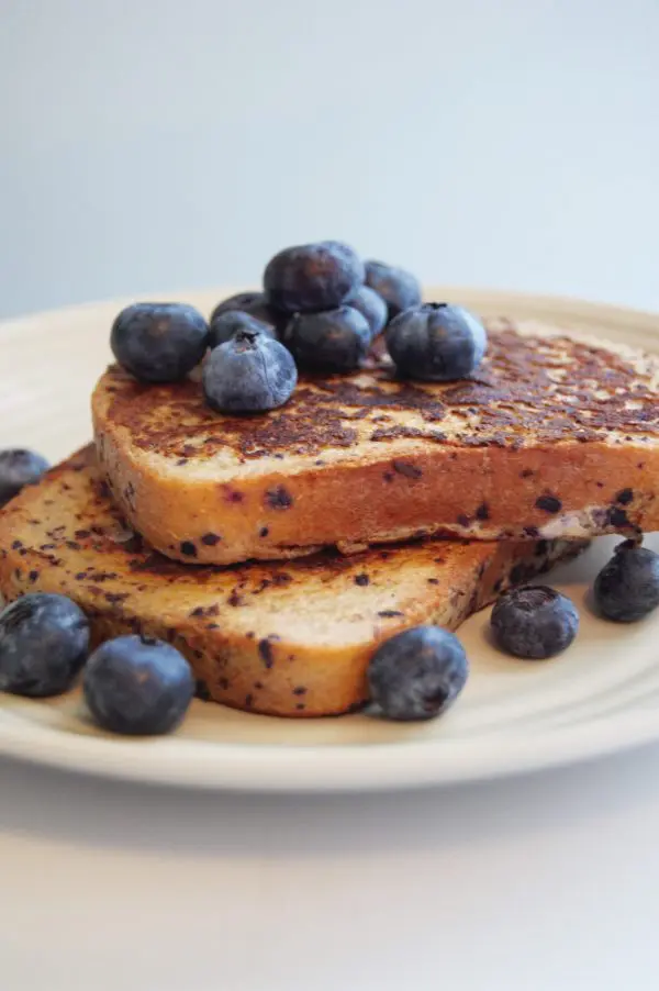 side, close up view of 2 slices of blueberry french toast on a beige plate topped with fresh blueberries.