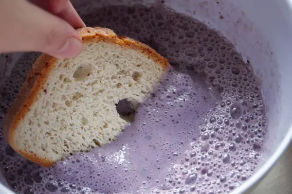 A piece of bread being dipped into a white bowl full of a purple-colored blueberry egg mixture to make blueberry French toast.