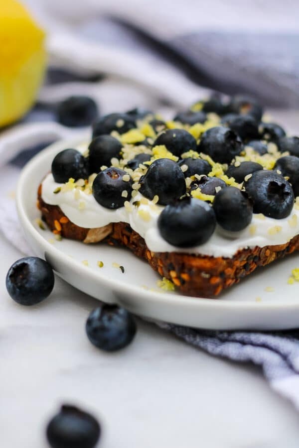 A close up of blueberry toast - crunchy whole grain and seed toast topped with creamy Greek yogurt, fresh blueberries, lemon zest, and hemp seeds.