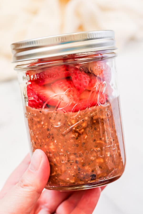 A wide mouth ball mason jar with chocolate protein overnight oats and fresh sliced strawberries held in a hand.