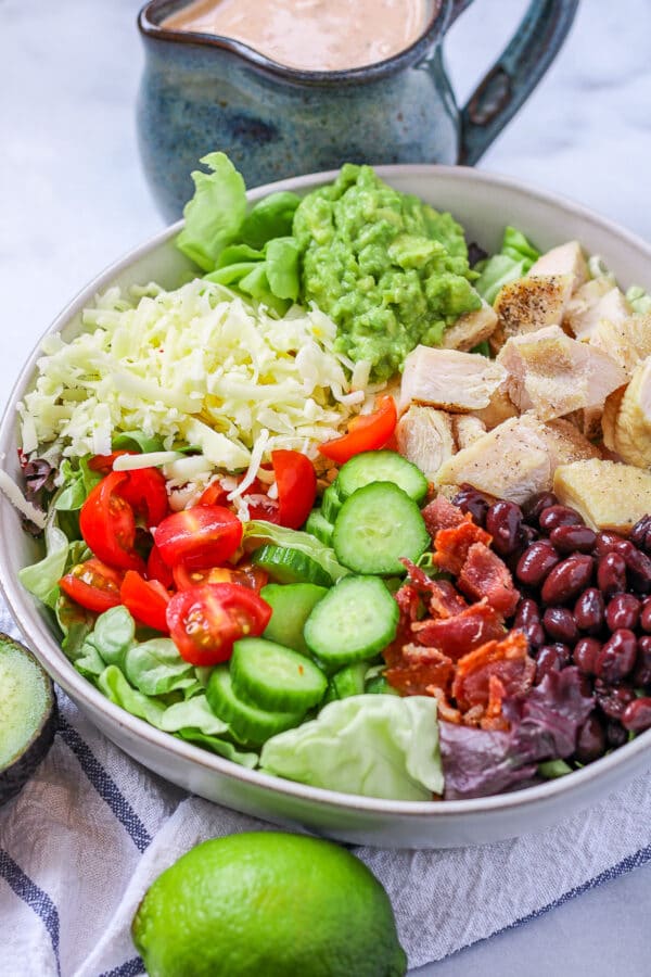 a close up of a southwest avocado salad, featuring a bed of lettuce topped with chicken, pepperjack cheese, guacamole, tomatoes, cucumbers, black beans, and bacon.