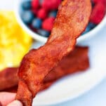 a slice of crisp turkey bacon being held up over a plate of eggs and berries