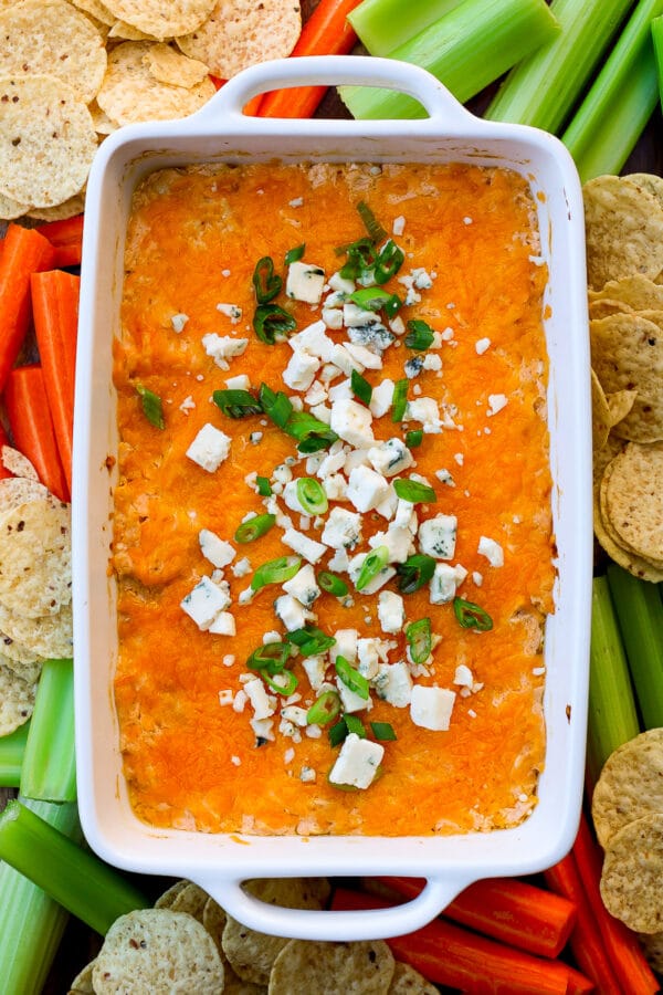 A white rectangular baking dish of healthy buffalo chicken dip topped with blue cheese crumbles and sliced green onion, surrounded by tortilla chips, celery sticks, and carrot sticks.