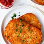 a white plate with two crispy hash brown patties next to a small dipping cup of ketchup