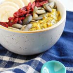 turmeric overnight oats in a white bowl topped with banana, pumpkin seeds, and goji berries