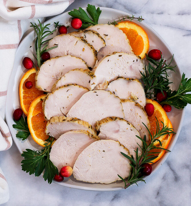 a platter of sliced, roasted dutch oven boneless turkey breast garnished with fresh herbs, orange slices, and fresh cranberries