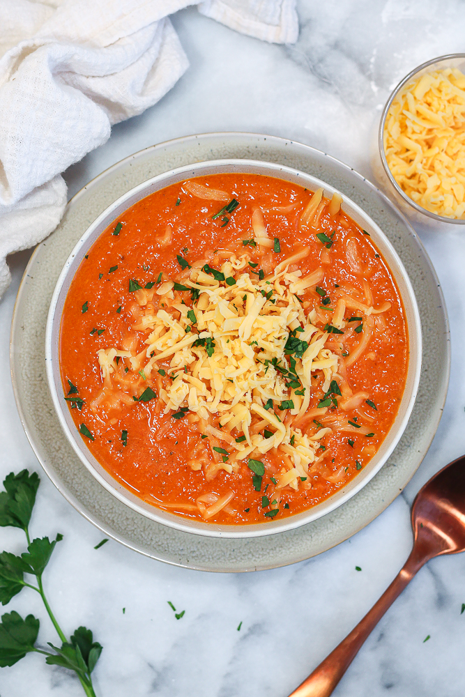 a bowl of roasted red pepper and gouda soup garnished with a pile of shredded gouda cheese and fresh parsley