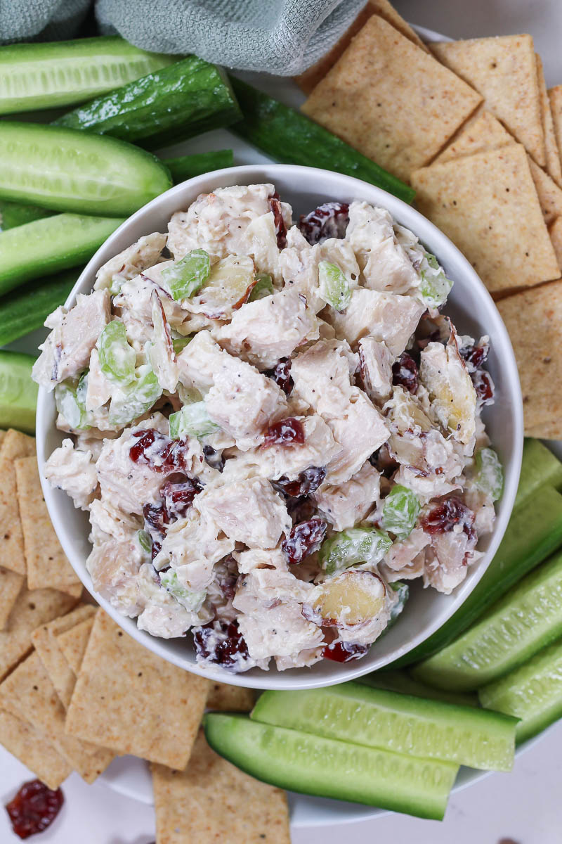 a bowl of cranberry chicken salad surrounded by crackers and cucumber slices
